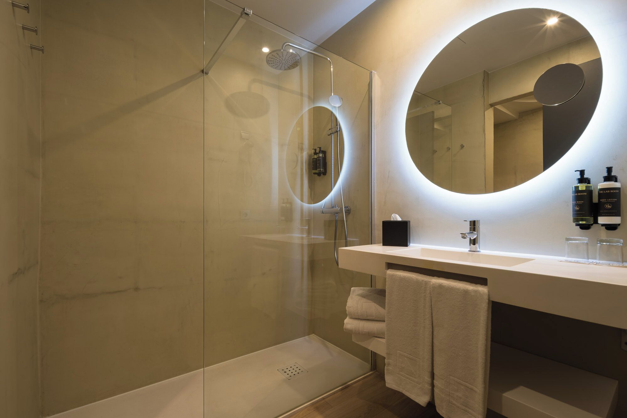 A modern bathroom with a glass-enclosed shower, a round mirror, and a white vanity with toiletries.