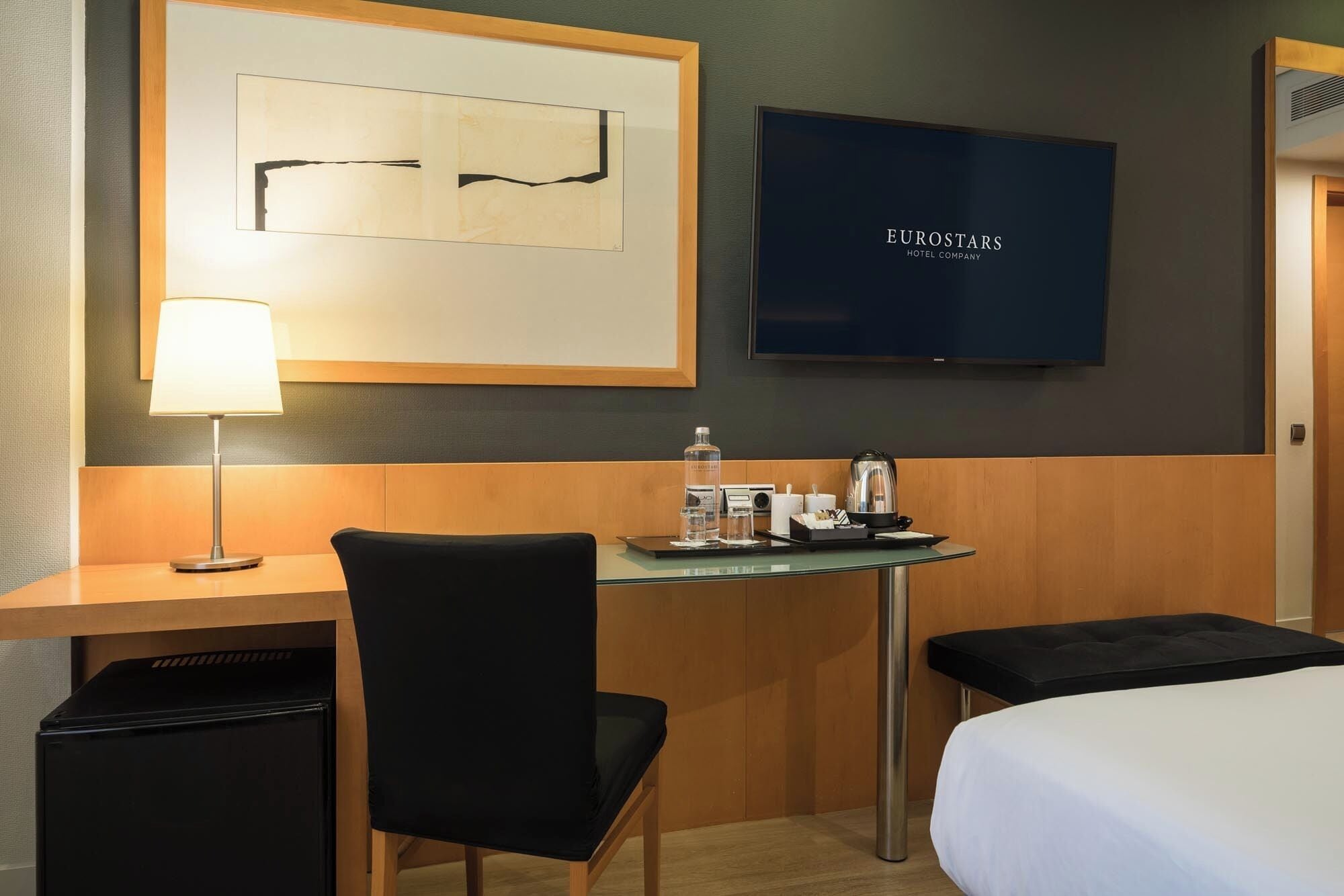 Una habitación de hotel con una cama, un escritorio con lámpara, una silla, una mesita con hervidor y botella, y un televisor en la pared.