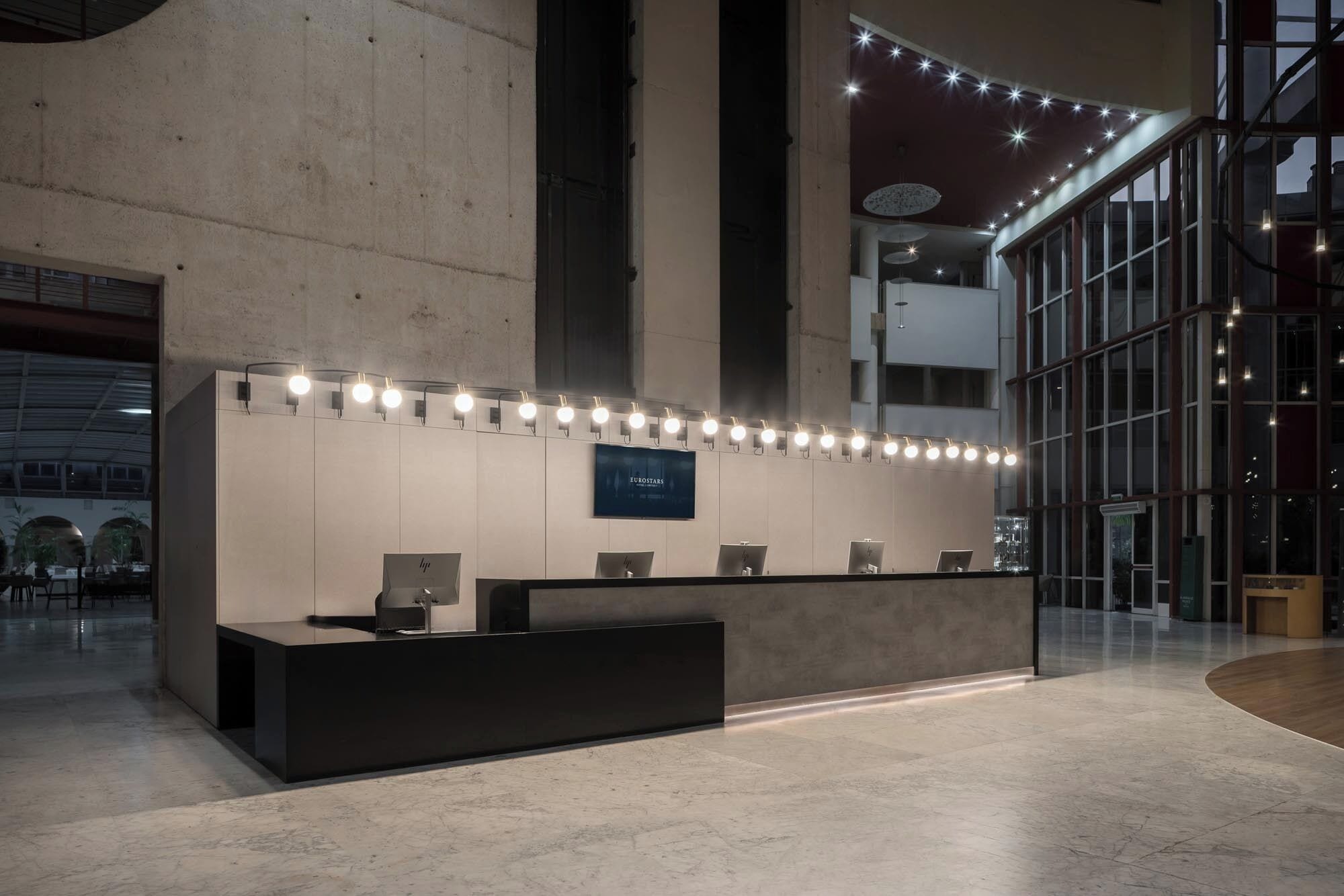 Una moderna recepción con pantalla digital, rodeada de luces, en un amplio vestíbulo con techos altos y grandes ventanales.