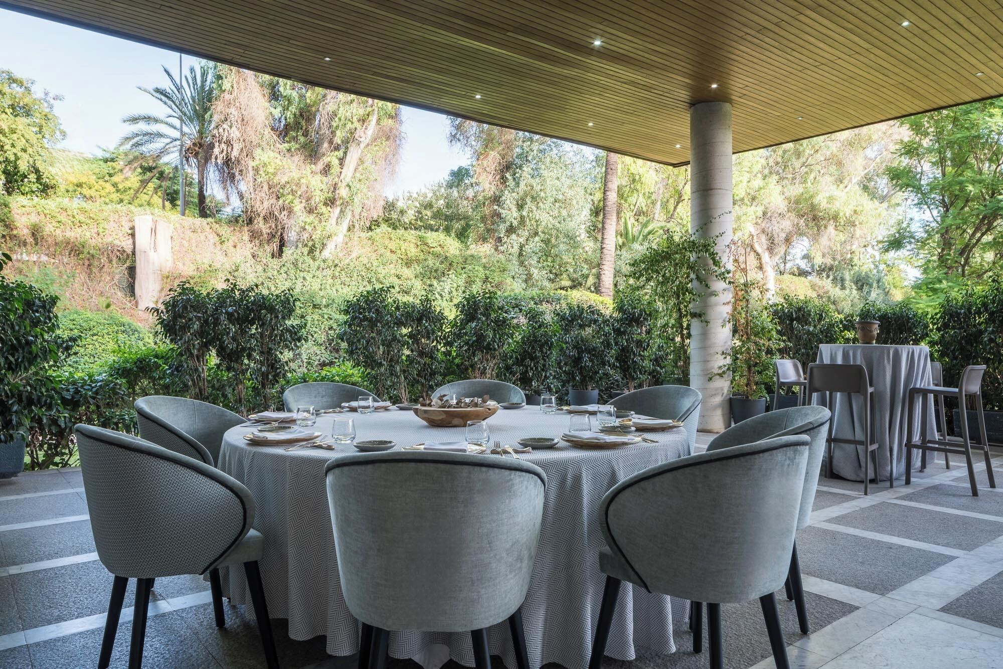 Un espacio exterior cubierto para comer con una mesa dispuesta para seis personas, sillas alrededor y vistas a un exuberante jardín.