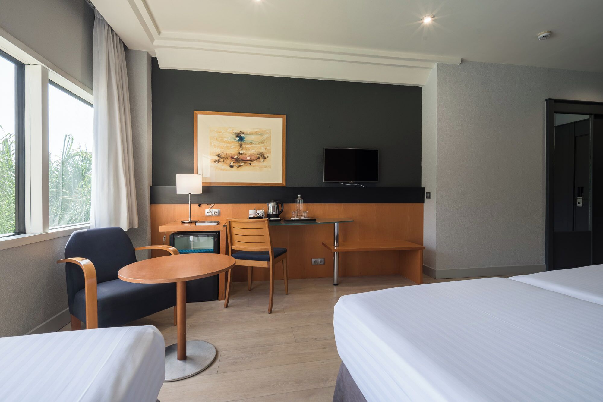 Habitación de hotel con dos camas, un escritorio con silla, una mesita, televisor montado en la pared y una ventana con cortinas.