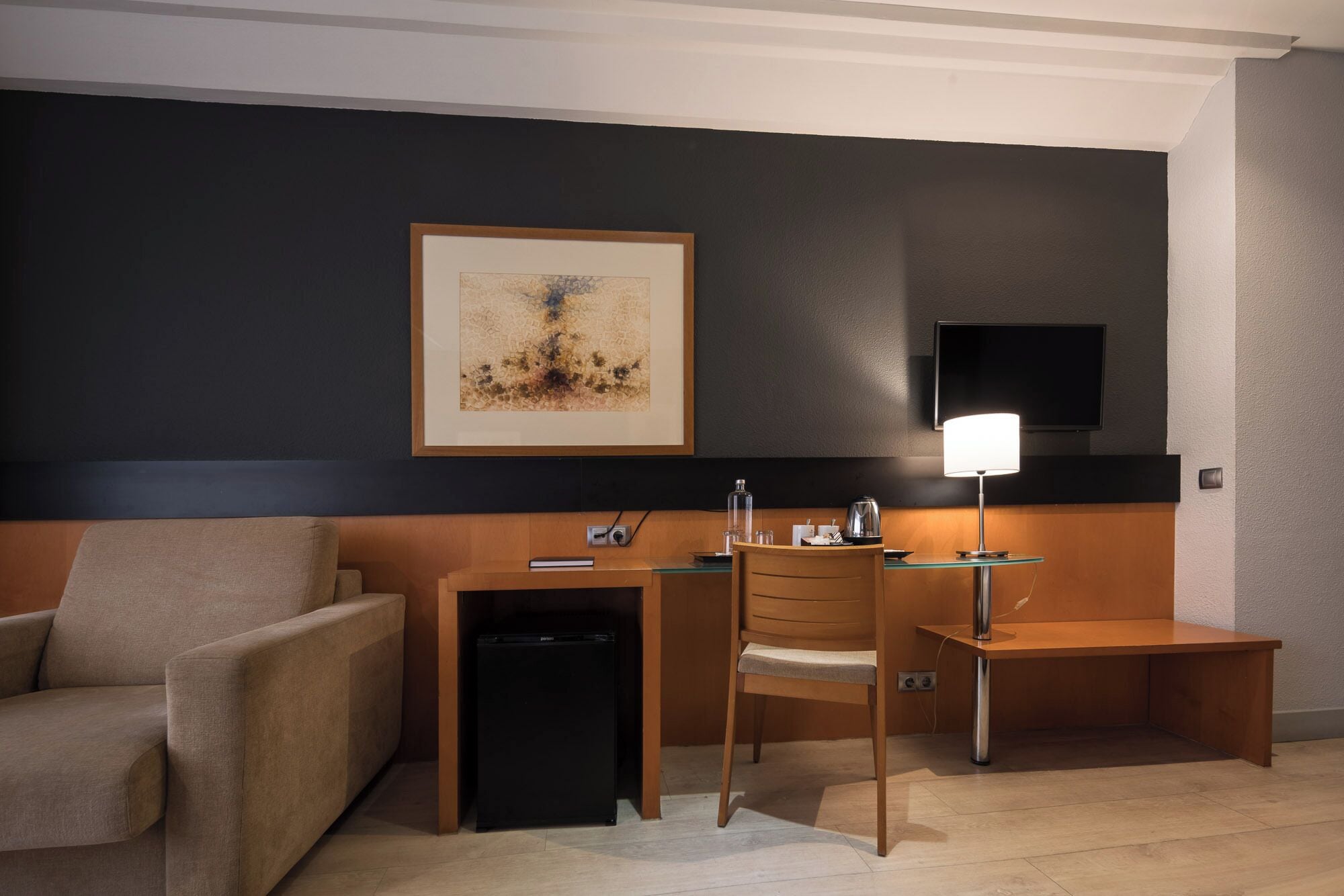 Habitación de hotel con escritorio, silla y una mesita con lámpara. Hay un televisor montado en la pared y una obra de arte enmarcada sobre el escritorio.