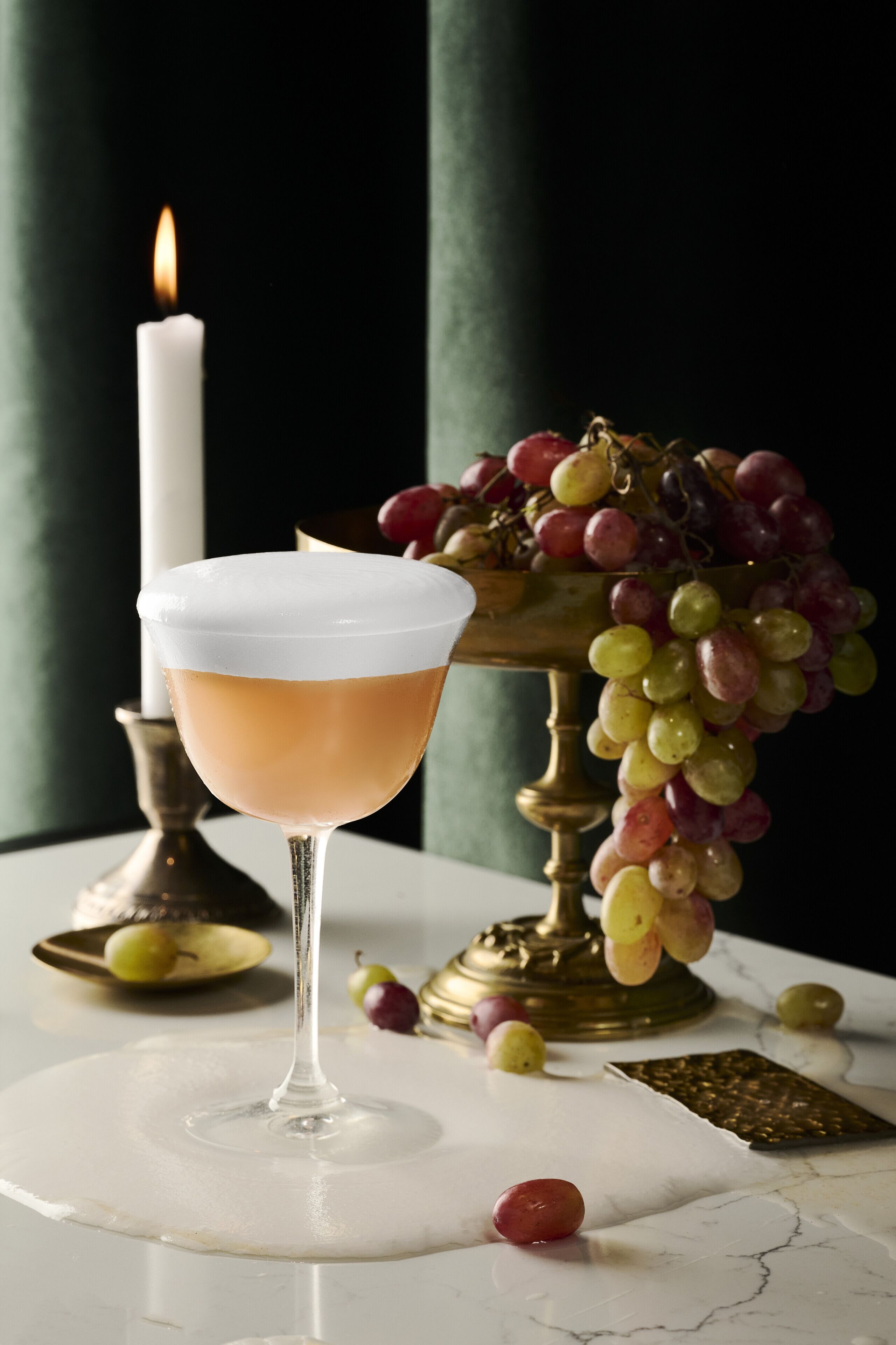 Un verre à cocktail à bordure blanche, une bougie allumée et un bol de raisins sur une surface en marbre.
