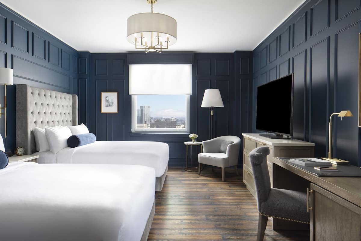 A hotel room with two beds, a desk, a chair, a TV, and a window with a city view.