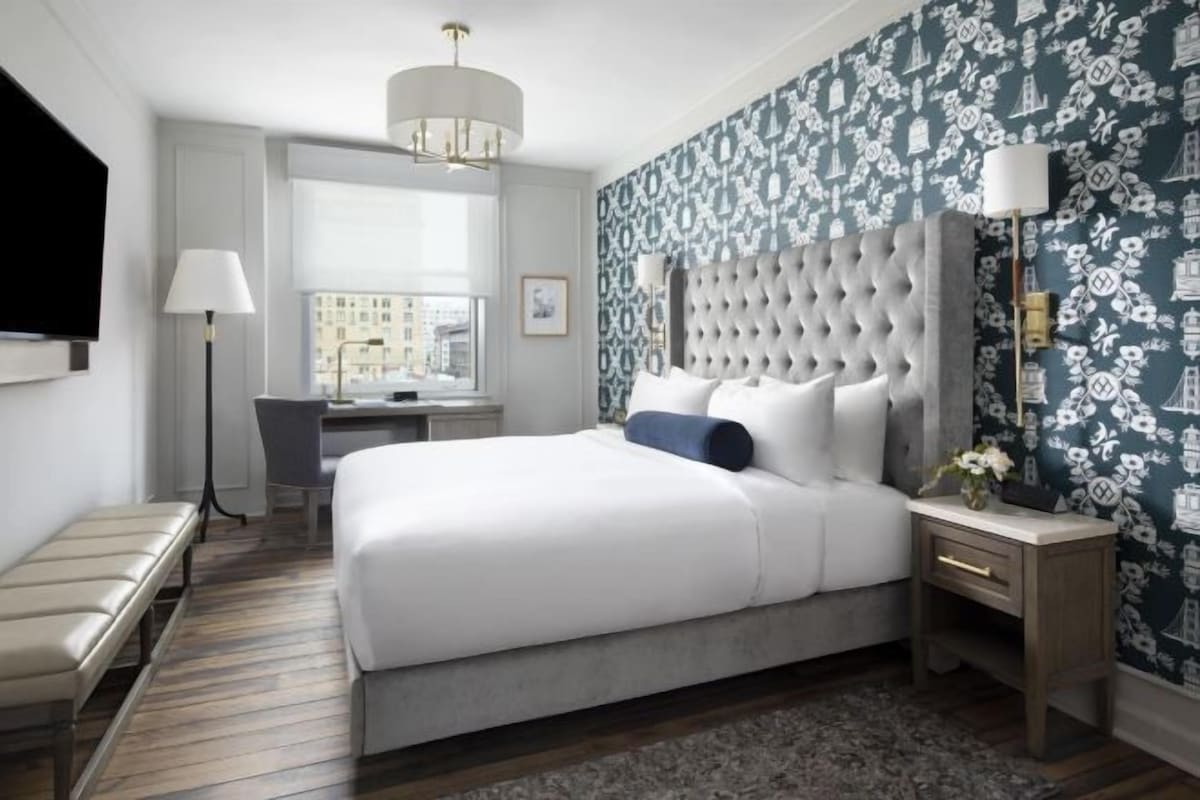 Een hotelkamer met een groot bed, een bureau, een stoel, een bankje, een televisie en behangpapier met een patroon.