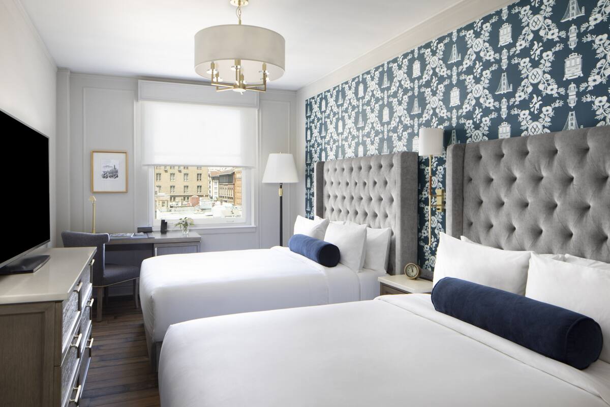 A hotel room with two beds, a desk, a TV, and a window with a city view.