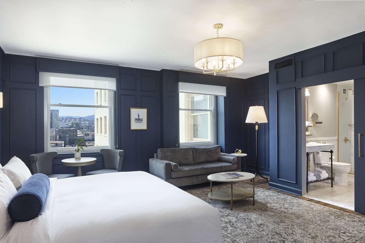 A hotel room with a large bed, a sofa, a small table, and a view of the city.