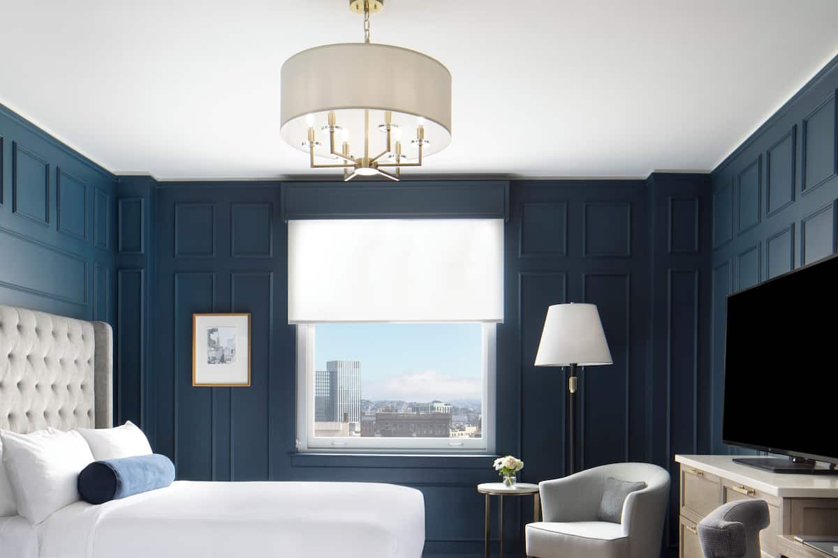 A hotel room with a large bed, a desk with a television, a chair, a lamp, and a window with a city view.