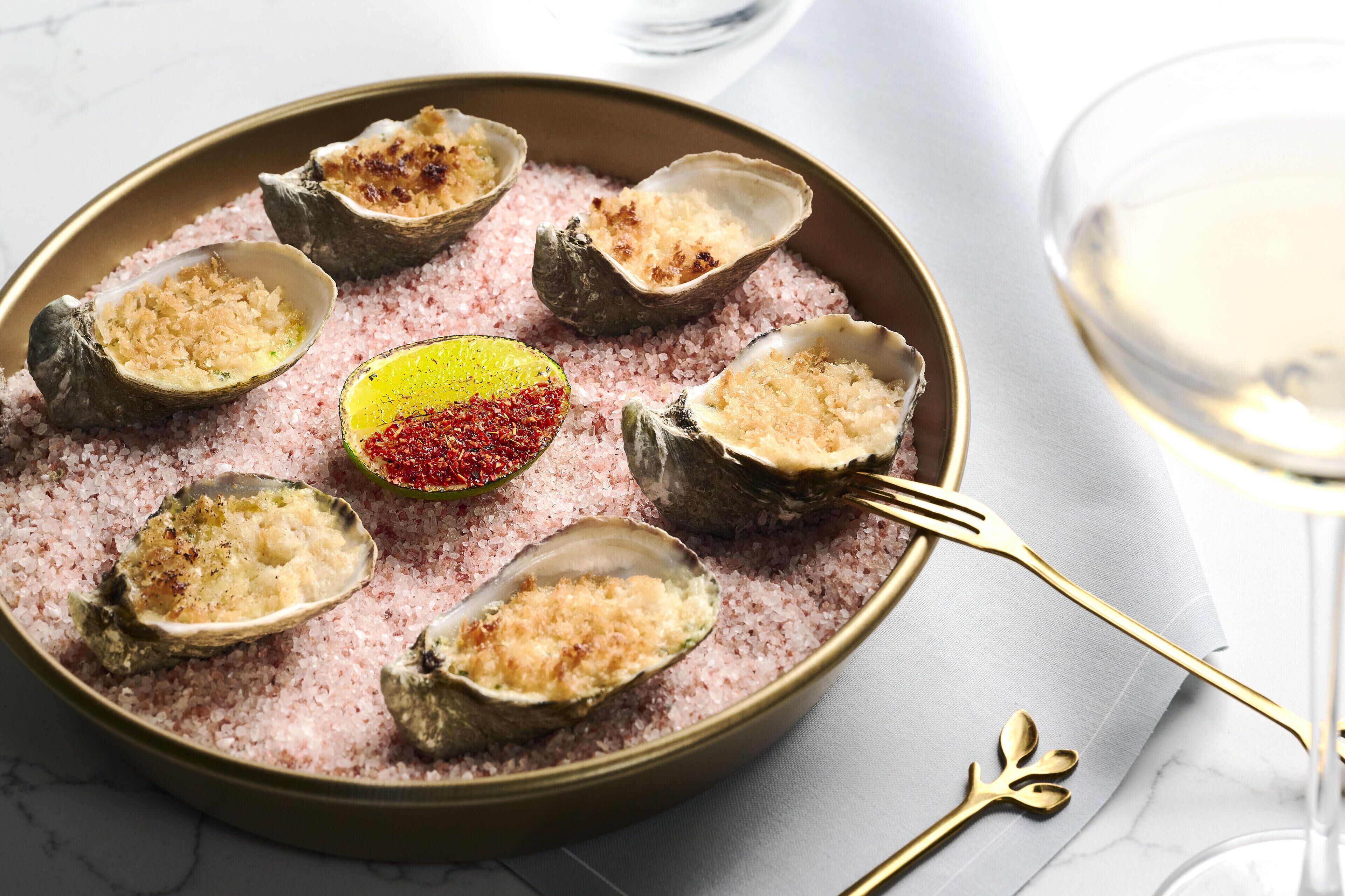 A plate of oysters on the half shell with a pink salt bed, a lemon half, and a red pepper flake.
