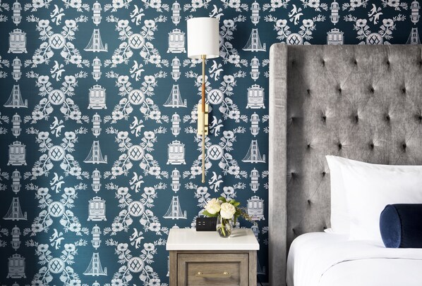 A bedroom with a tufted headboard, a bedside table with a vase of flowers, and a wall with a patterned wallpaper featuring white illustrations of buildings and flowers. - Beacon Grand, A Union Square Hotel  (San Francisco)