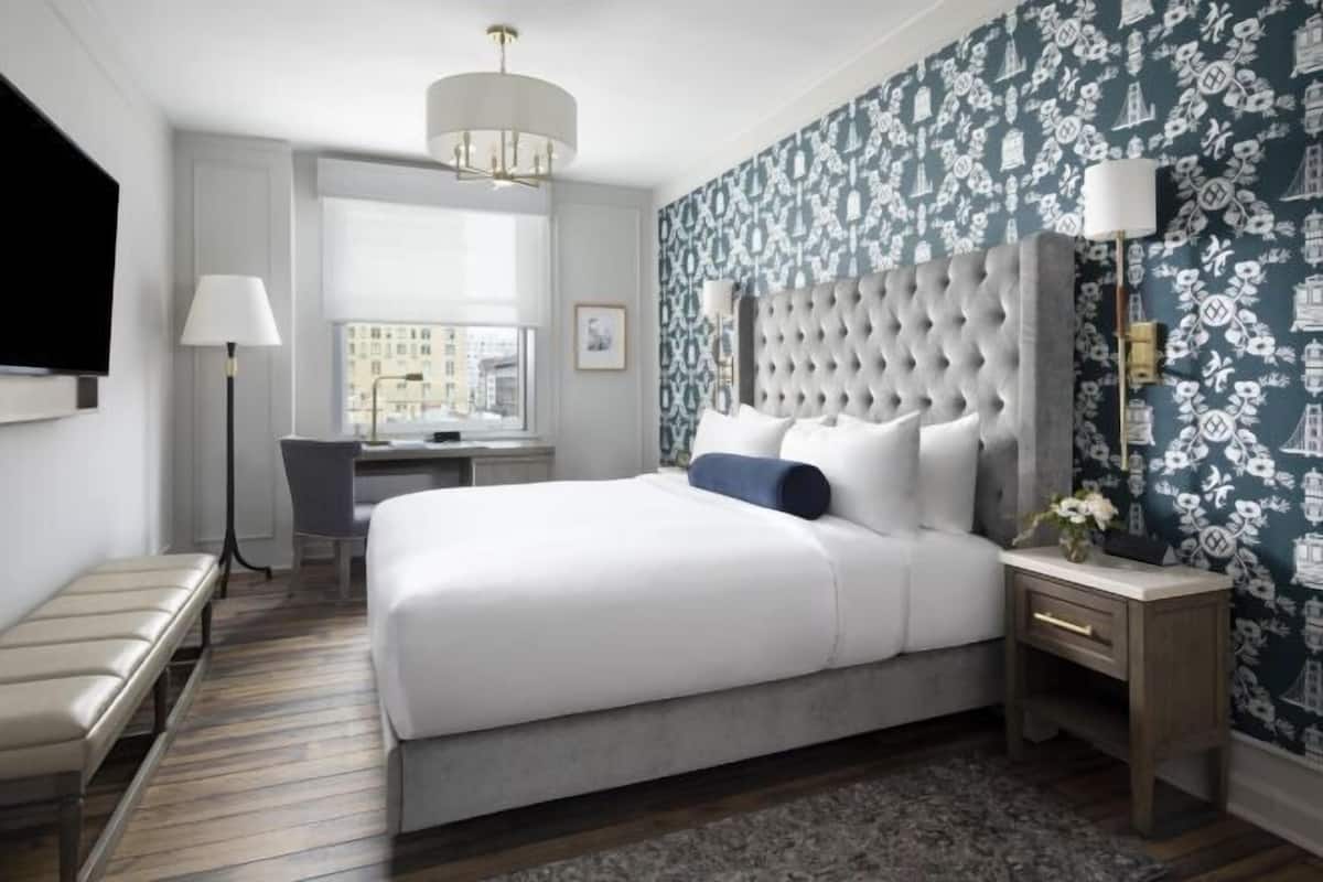 A hotel room with a large bed, a desk, a chair, a bench, a TV, and a patterned wallpaper.