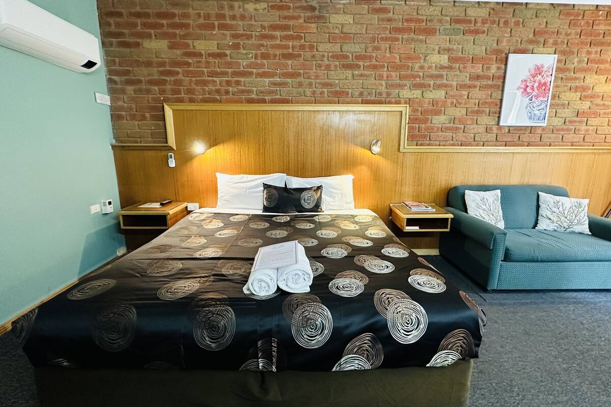 A hotel room with a large bed, a wooden headboard, a green armchair, and a brick wall.