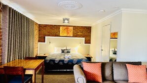 A hotel room with a brick accent wall, a bed with a patterned bedspread, a wooden desk, a sofa with patterned cushions, and a ceiling light fixture. - Early Australian Motor Inn (Mildura)