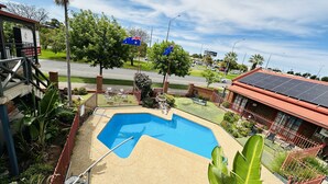 A swimming pool with a diving board, surrounded by a fenced garden with palm trees and a view of a street with cars and buildings. - Early Australian Motor Inn (Mildura)