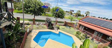 A swimming pool with a diving board, surrounded by a fenced garden with palm trees and a view of a street with cars and buildings.