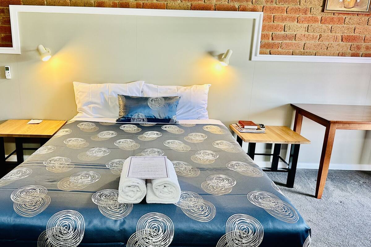 A neatly made bed with a patterned bedspread, two bedside tables, a nightstand, and a small wooden table with a book and remote control.
