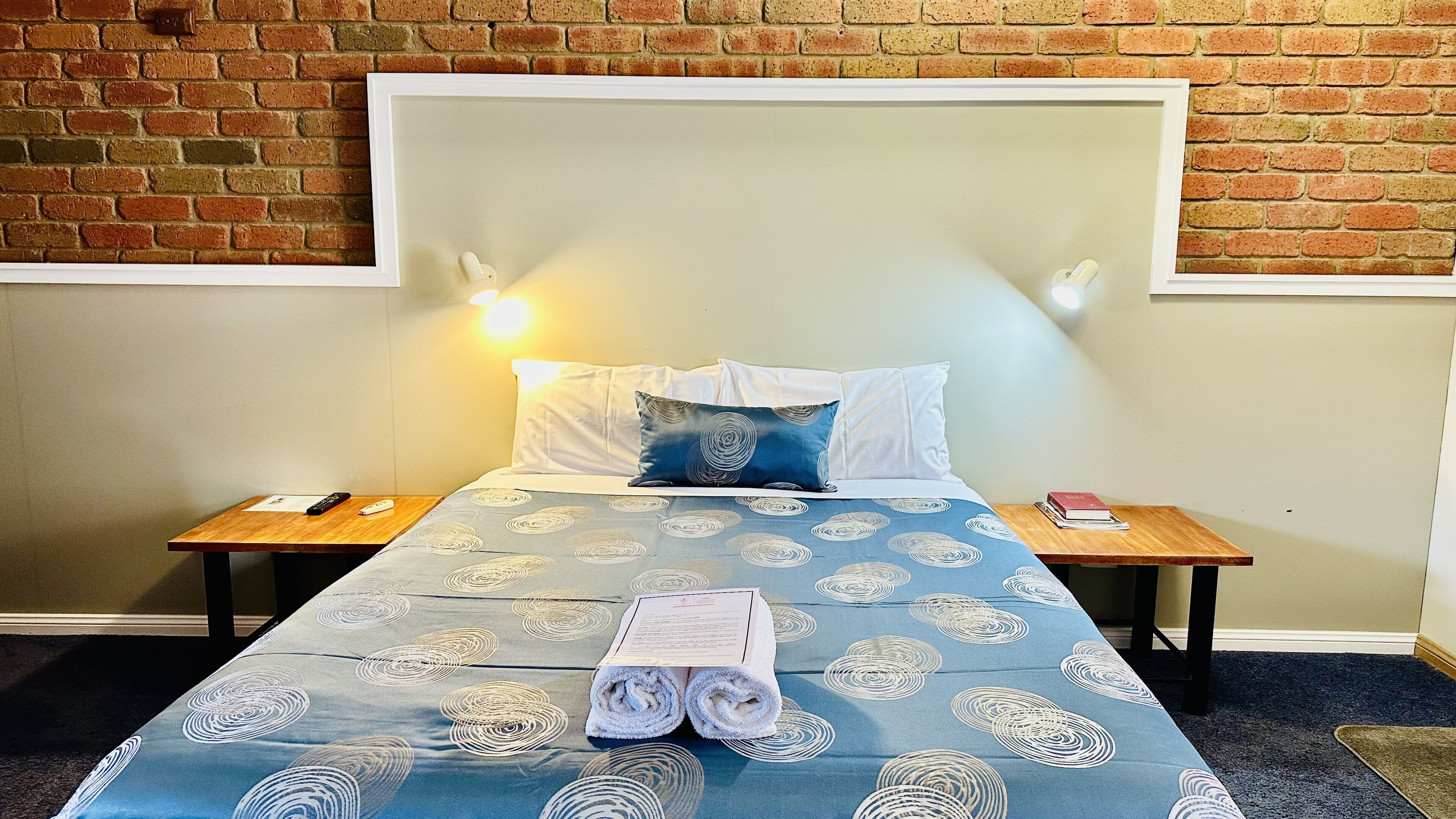 Cama bem arrumada com colcha azul e branca estampada, dois travesseiros e um par de toalhas enroladas. Duas mesas de cabeceira, uma com um controle remoto e um livro, e outra com uma luminária. O quarto tem uma parede de tijolos e uma cabeceira branca.