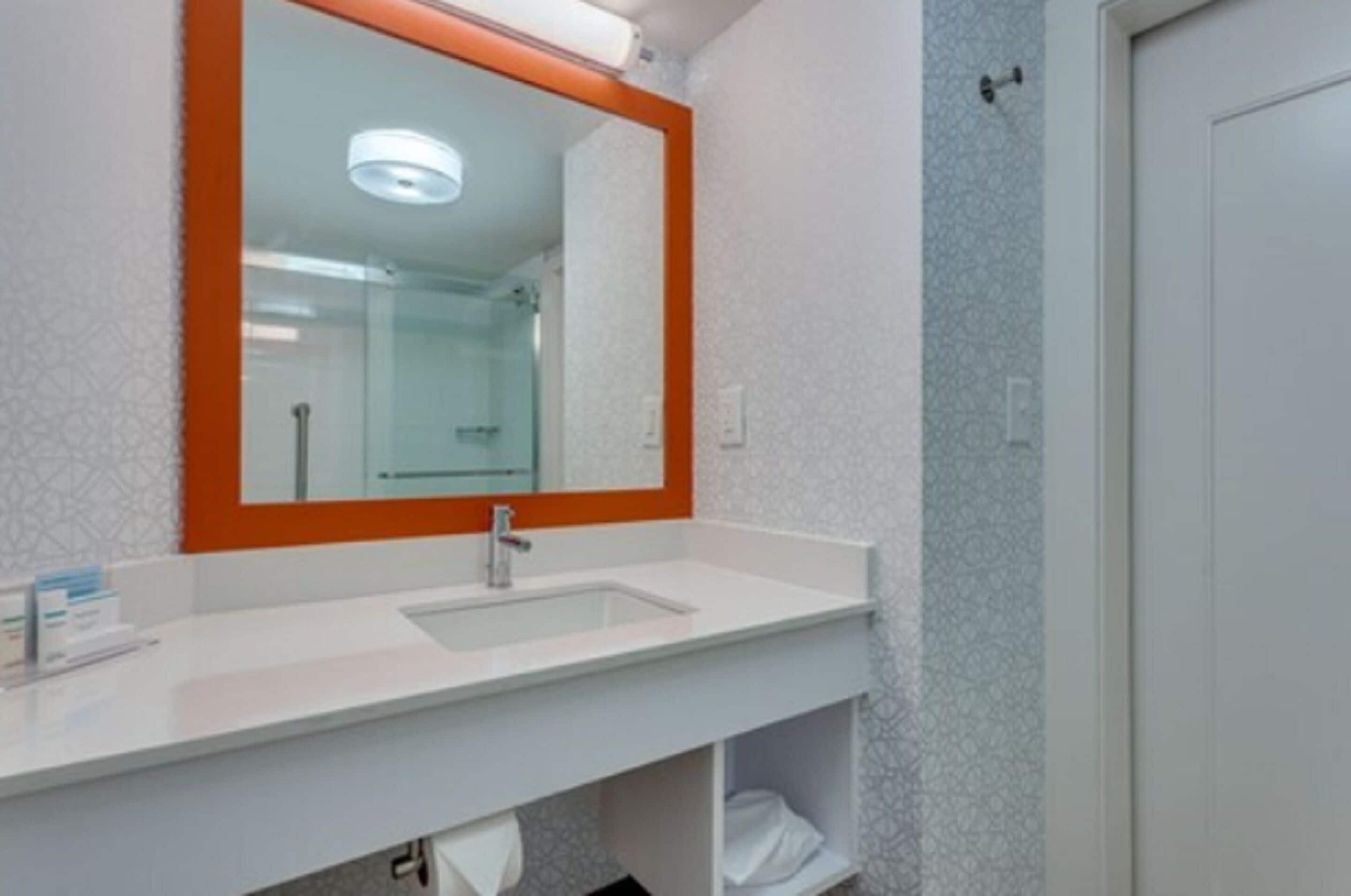 Un baño con un lavamanos blanco, un espejo con marco anaranjado y una ducha visible al fondo.