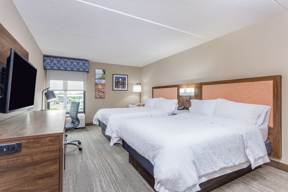 A hotel room with two beds, a desk with a TV, a chair, a window with a view, and a door.