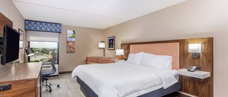 A hotel room with a bed, desk, chair, and a view of the city.