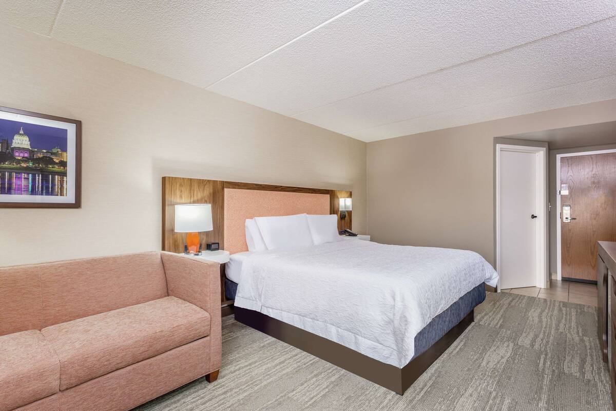 A hotel room with a bed, a nightstand, a lamp, a framed picture on the wall, and a door.