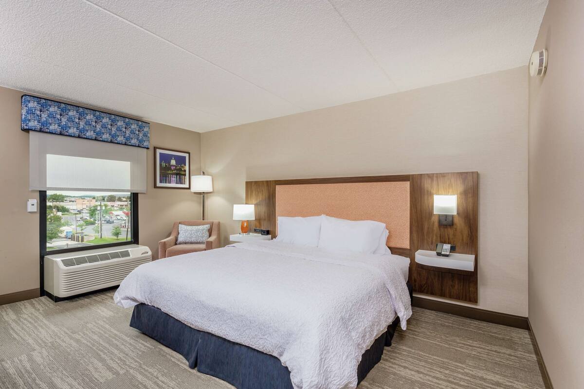 A hotel room with a large bed, a window with a city view, a desk with a lamp, and a wall-mounted phone.