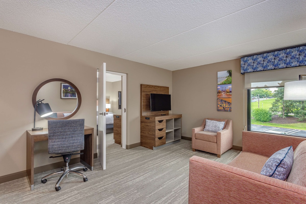 A hotel room with a wooden desk, a comfortable sofa, a television, and a large window offering a view of the outdoors.