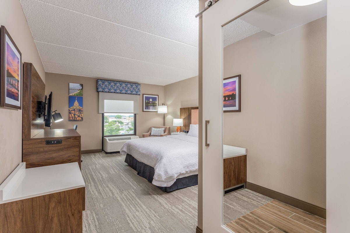 A hotel room with a large bed, a desk with a mirror, a dresser, and a window with a view of buildings.