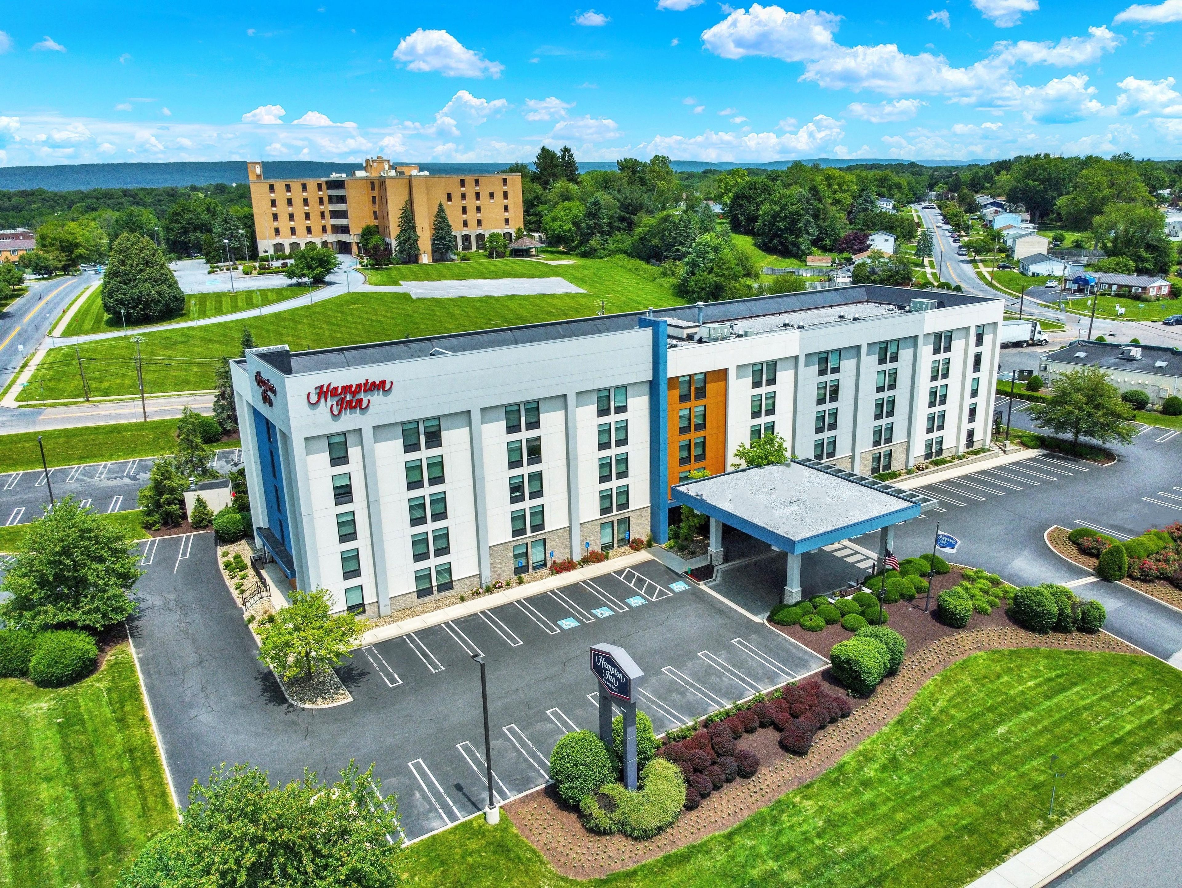 Um hotel Hampton Inn com estacionamento, área externa coberta e vista para um edifício maior ao fundo.