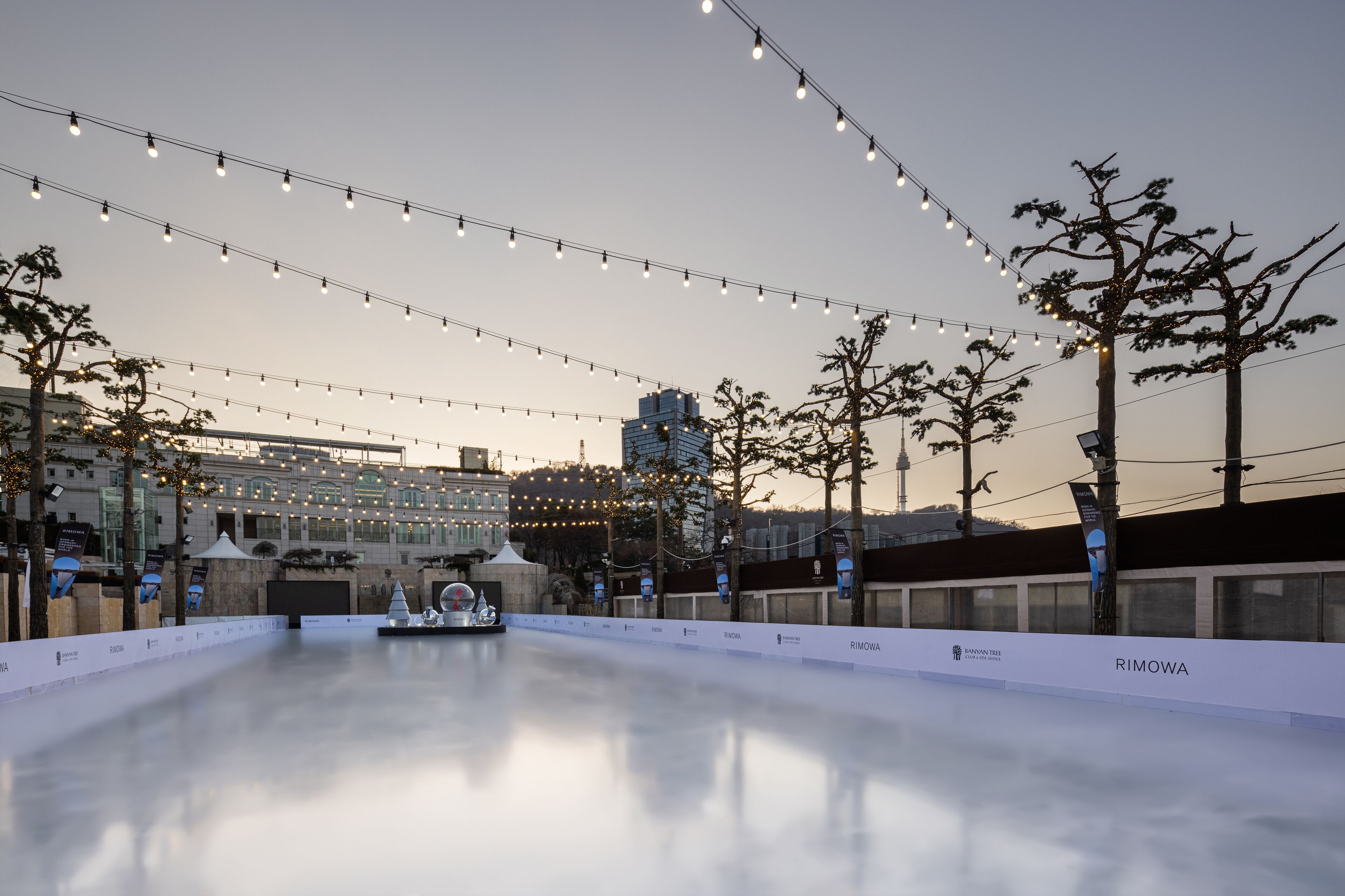 Una pista de hielo al aire libre con luces colgantes, árboles y edificios en el fondo.