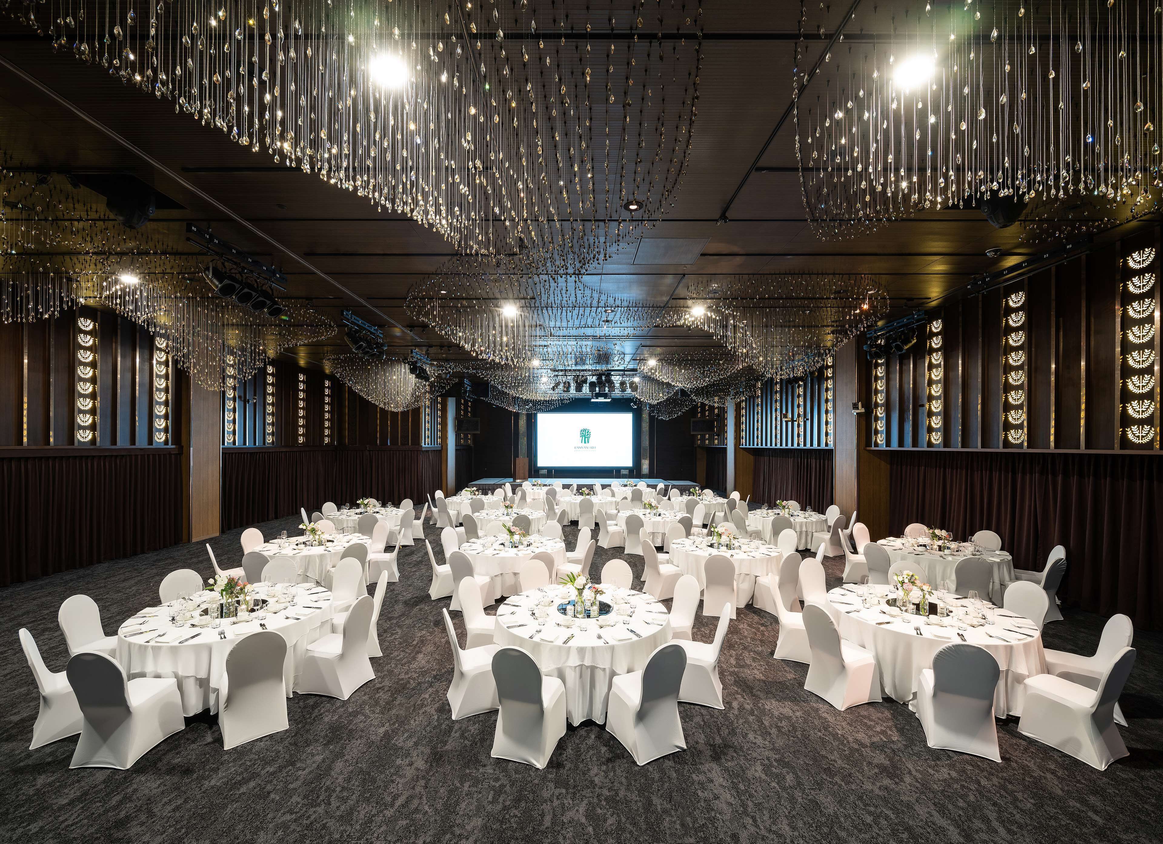 A banquet hall with round tables set for dining, a stage with a presentation screen, and decorative hanging lights.