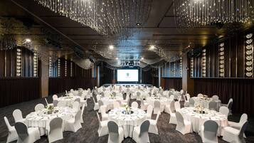 A banquet hall with round tables set for dining, a stage with a presentation screen, and decorative hanging lights.