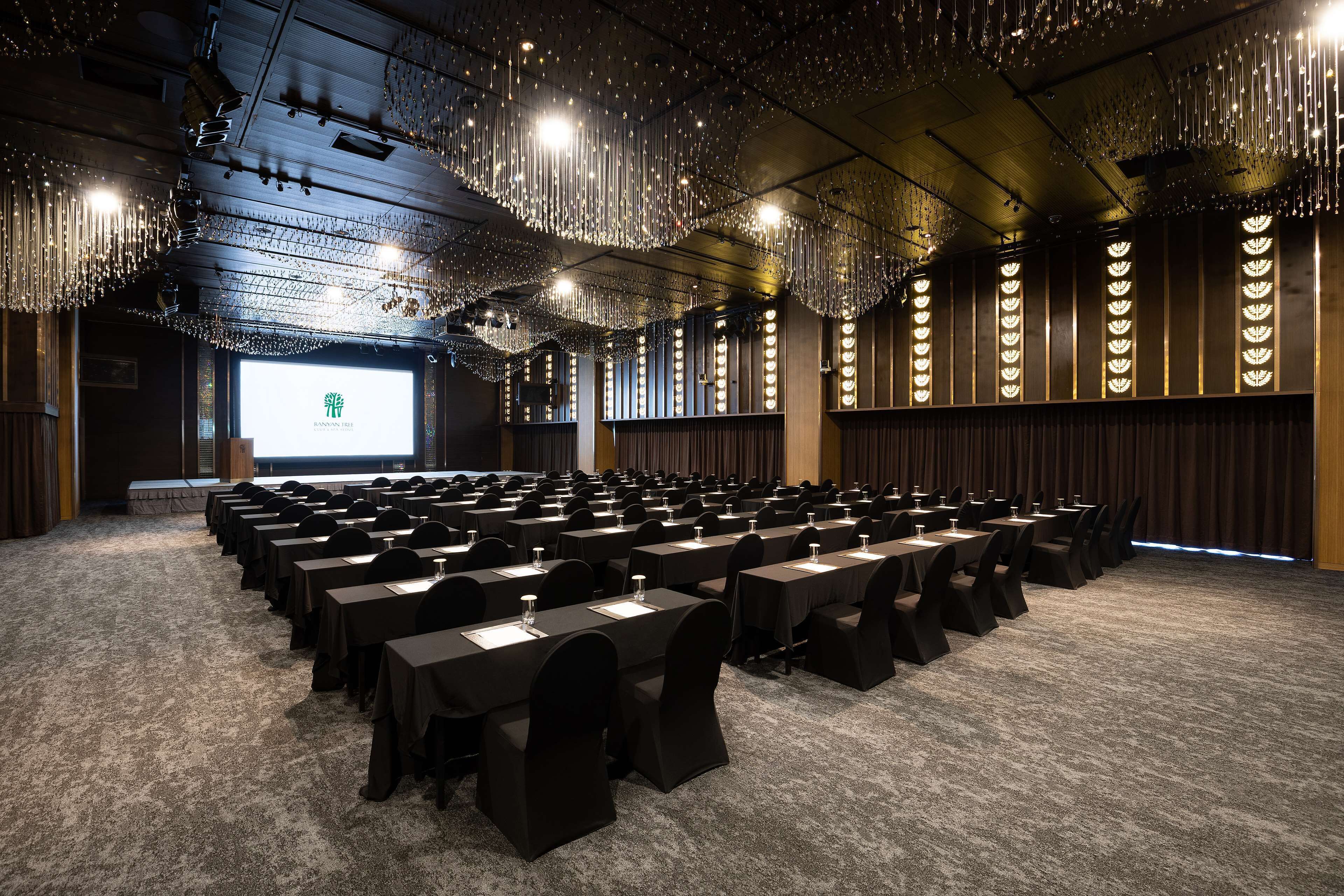 A conference room with rows of tables and chairs, a large screen at the front, and a ceiling with hanging lights.