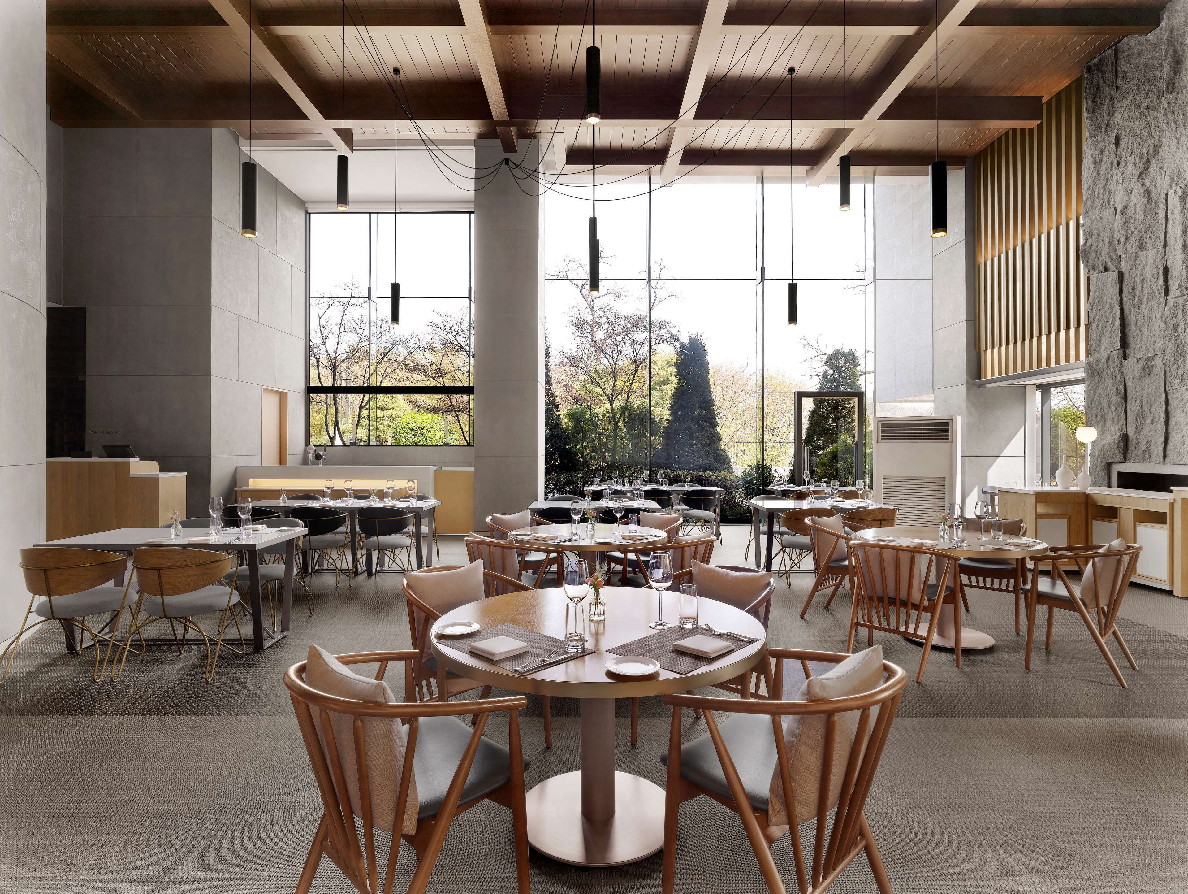 A modern dining area with round tables and wooden chairs, large windows offering a view of trees and a building, and a stone wall on one side.