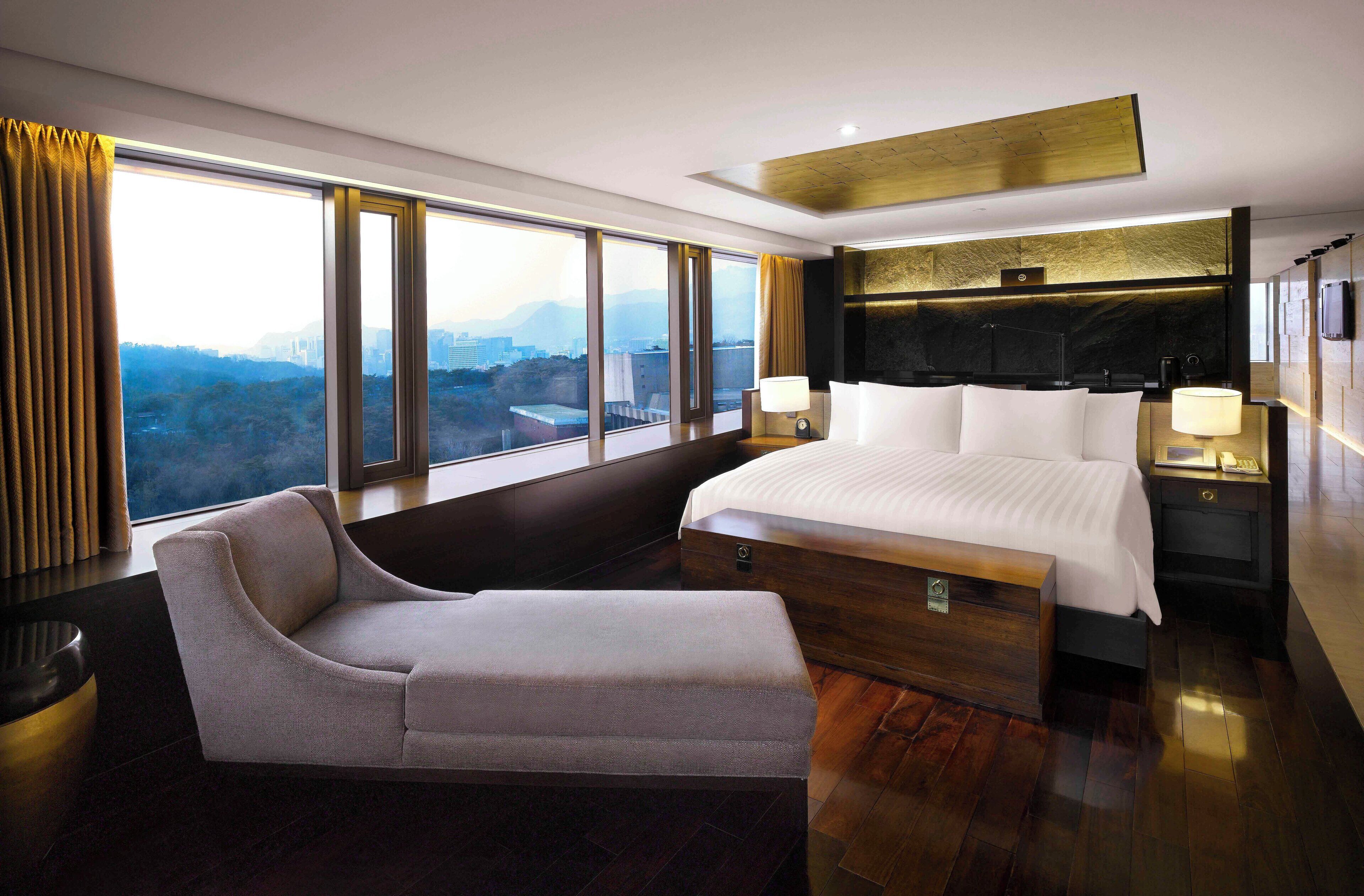 A modern hotel room with a large bed, a chaise lounge, a view of the city, and a wooden floor.