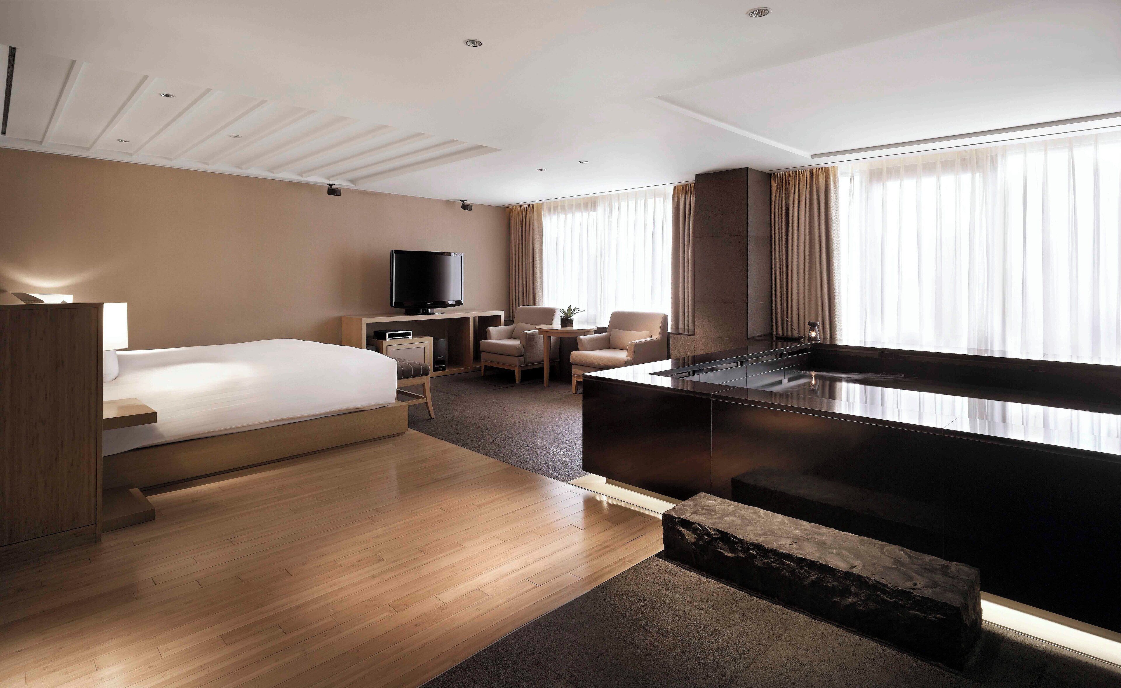 Una habitación de hotel moderna con una cama grande, un escritorio con televisor de pantalla plana, una zona de estar con dos sillones, una mesita y una bañera espaciosa.