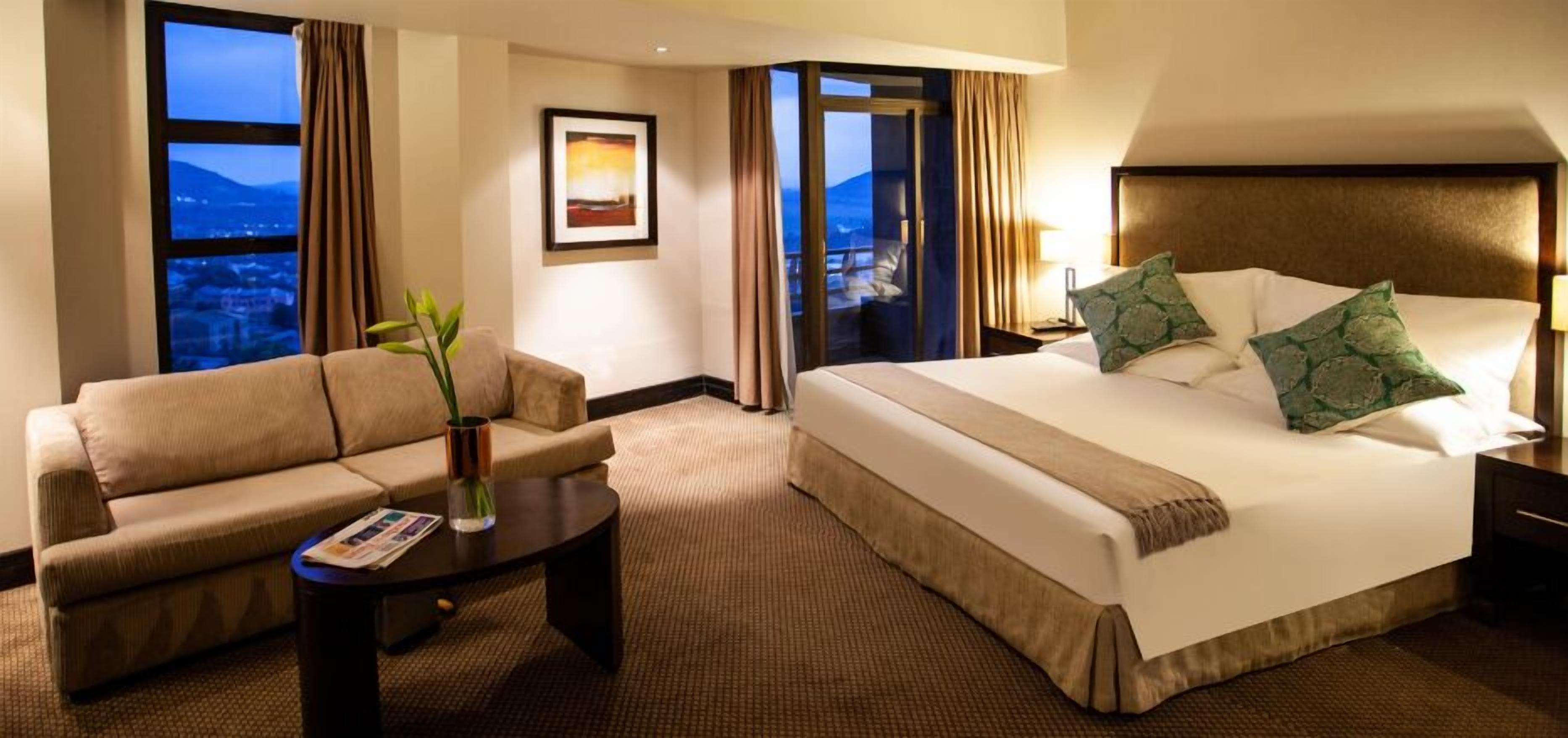 A hotel room with a bed, a sofa, a coffee table, and a view of the cityscape.