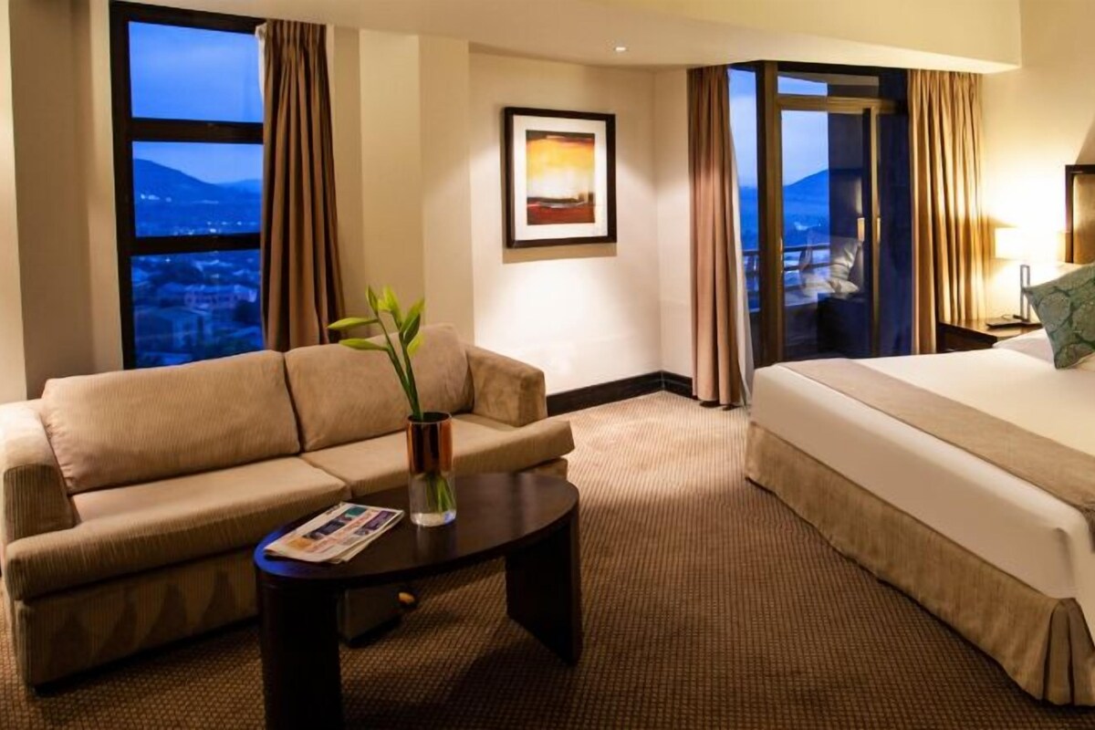 A hotel room with a bed, a sofa, a coffee table, and a view of the cityscape.