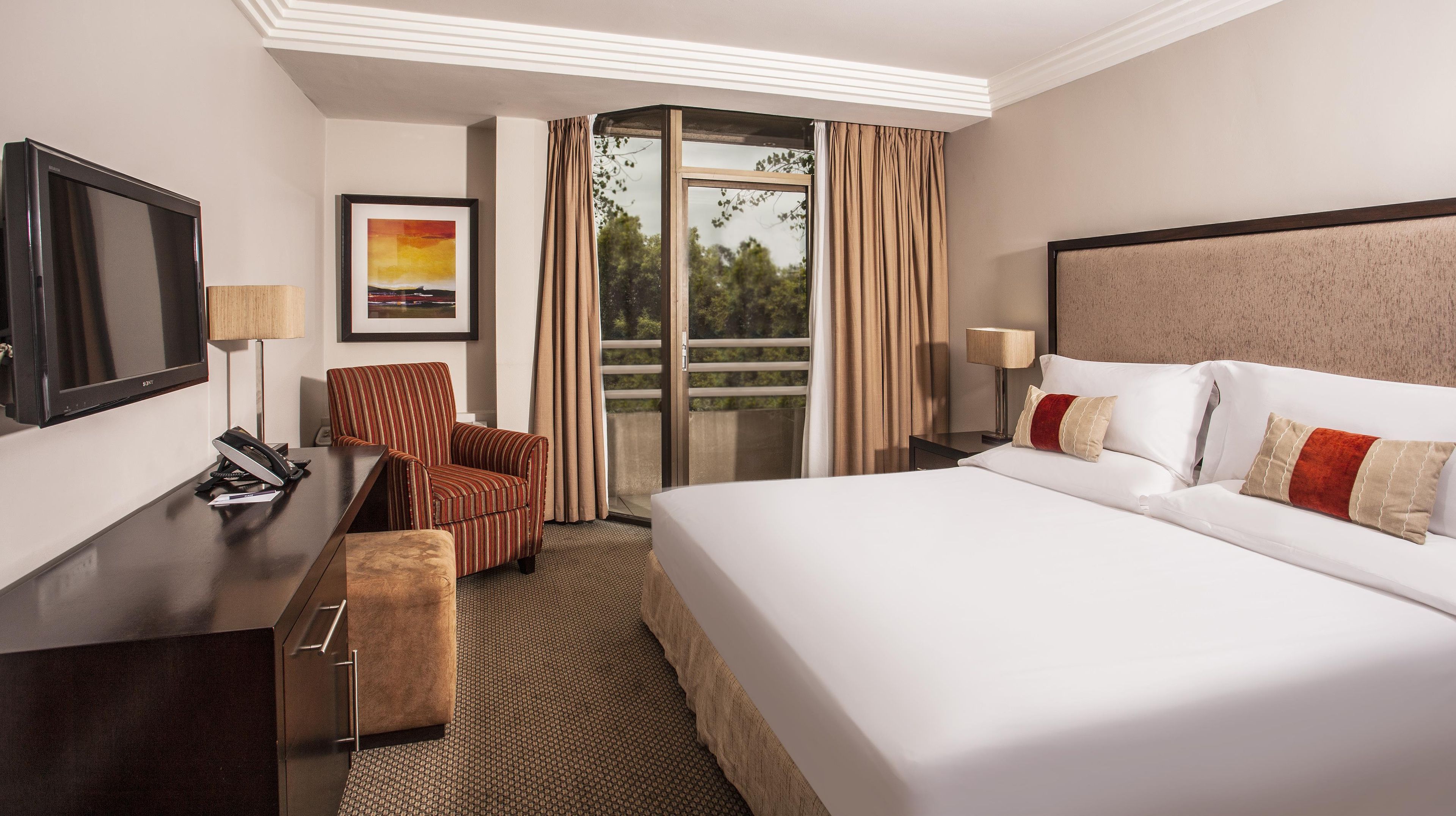 Quarto de hotel com cama, escrivaninha, cadeira, televisão e janela com cortinas.