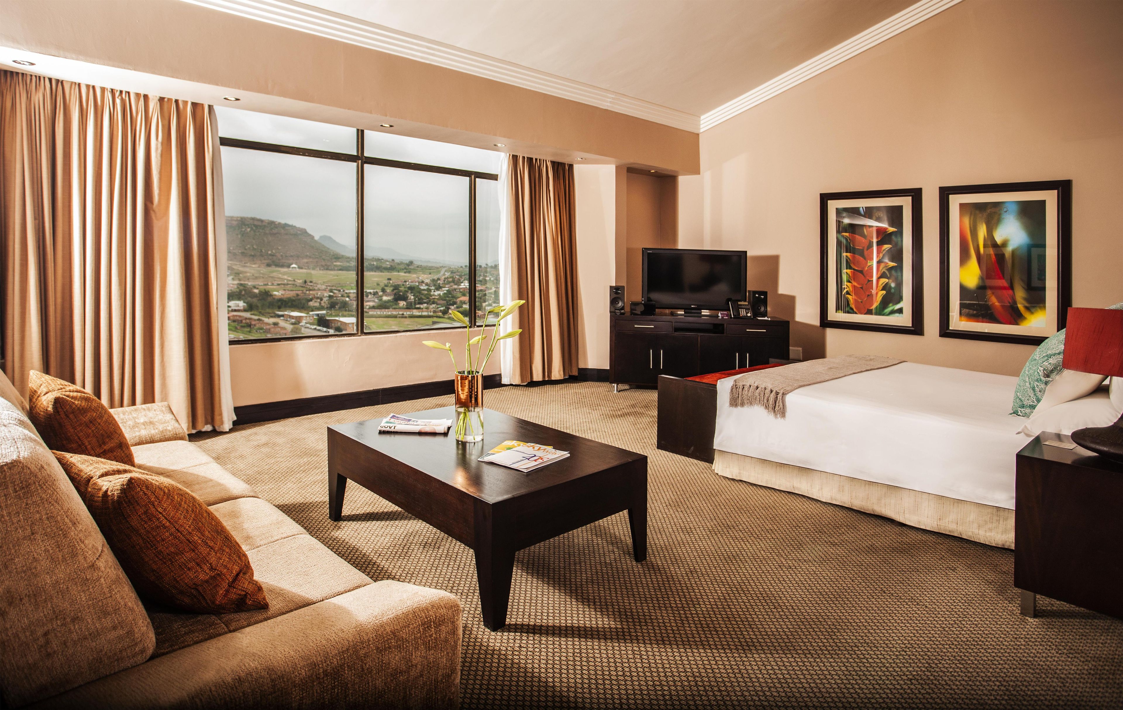 A hotel room with a large bed, a sofa, a coffee table, a TV, and a view of a landscape through the window.