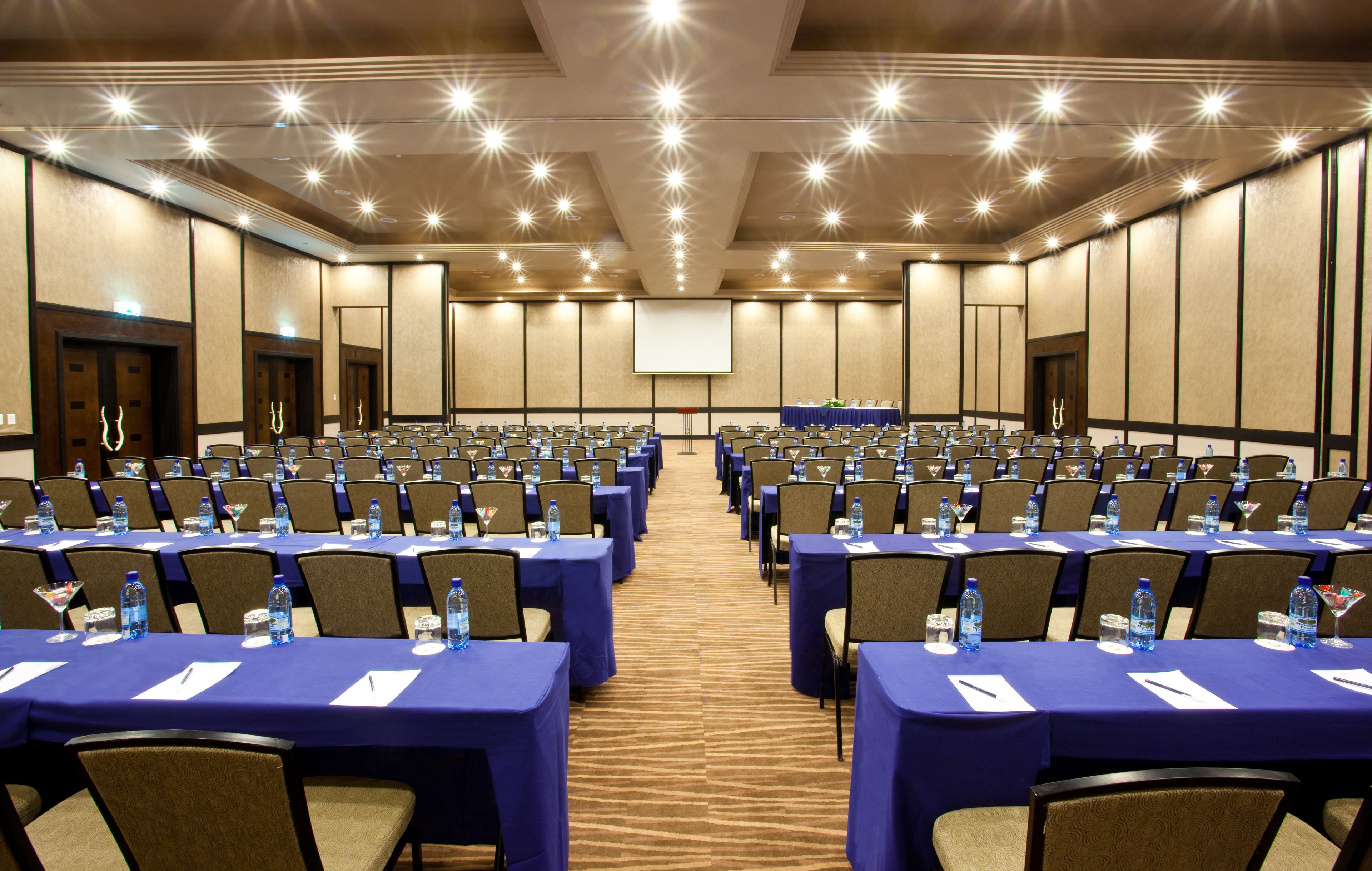 A conference room with rows of tables, chairs, and a projector screen at the front.