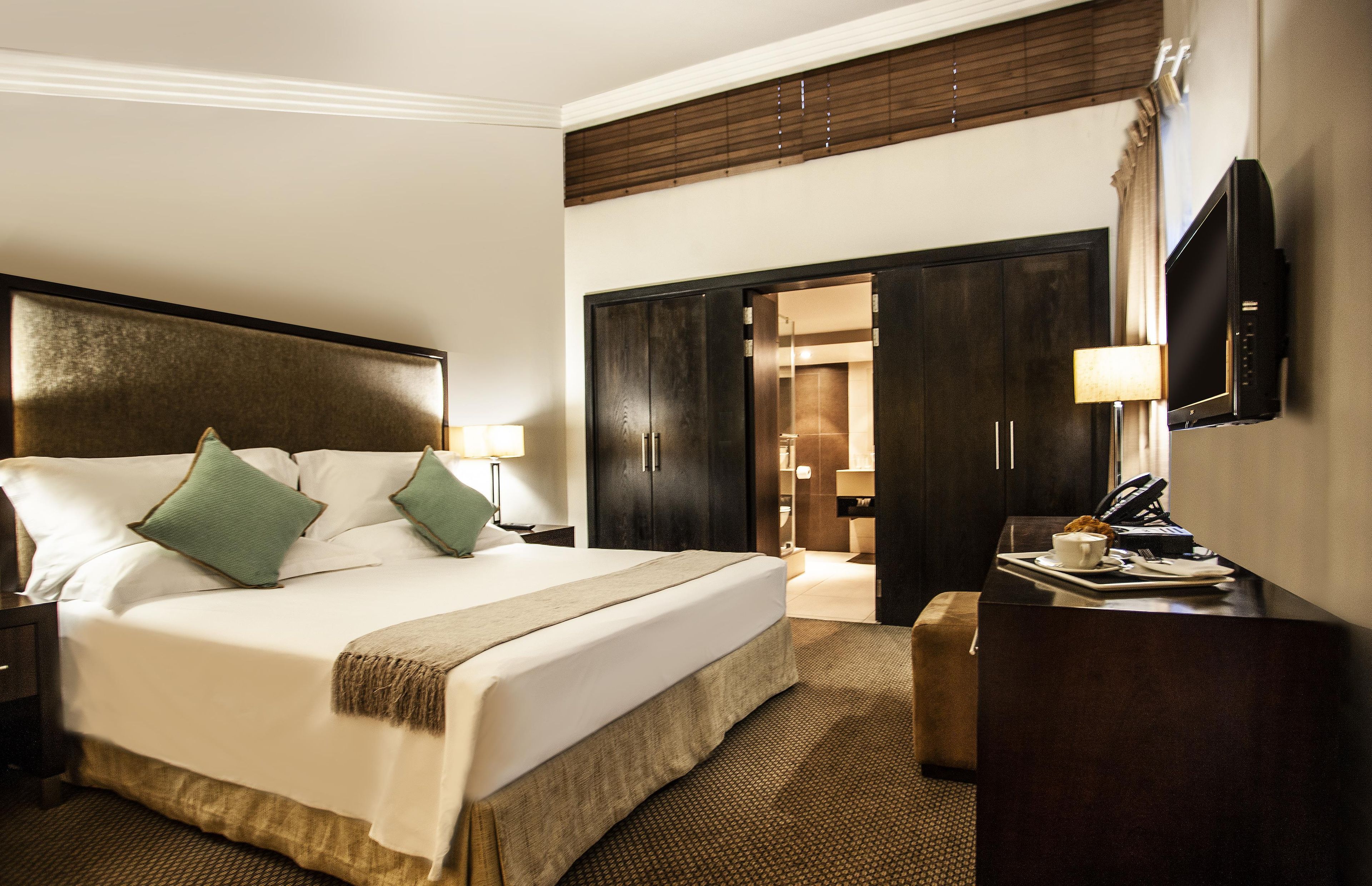 A hotel room with a large bed, a desk with a flat-screen TV, a wardrobe, and a bathroom visible through an open door.