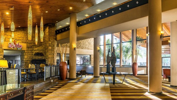 A spacious lobby with a stone wall, a bar area, and large windows offering a view of the outdoors. - Avani Lesotho Hotel & Casino (Maseru)