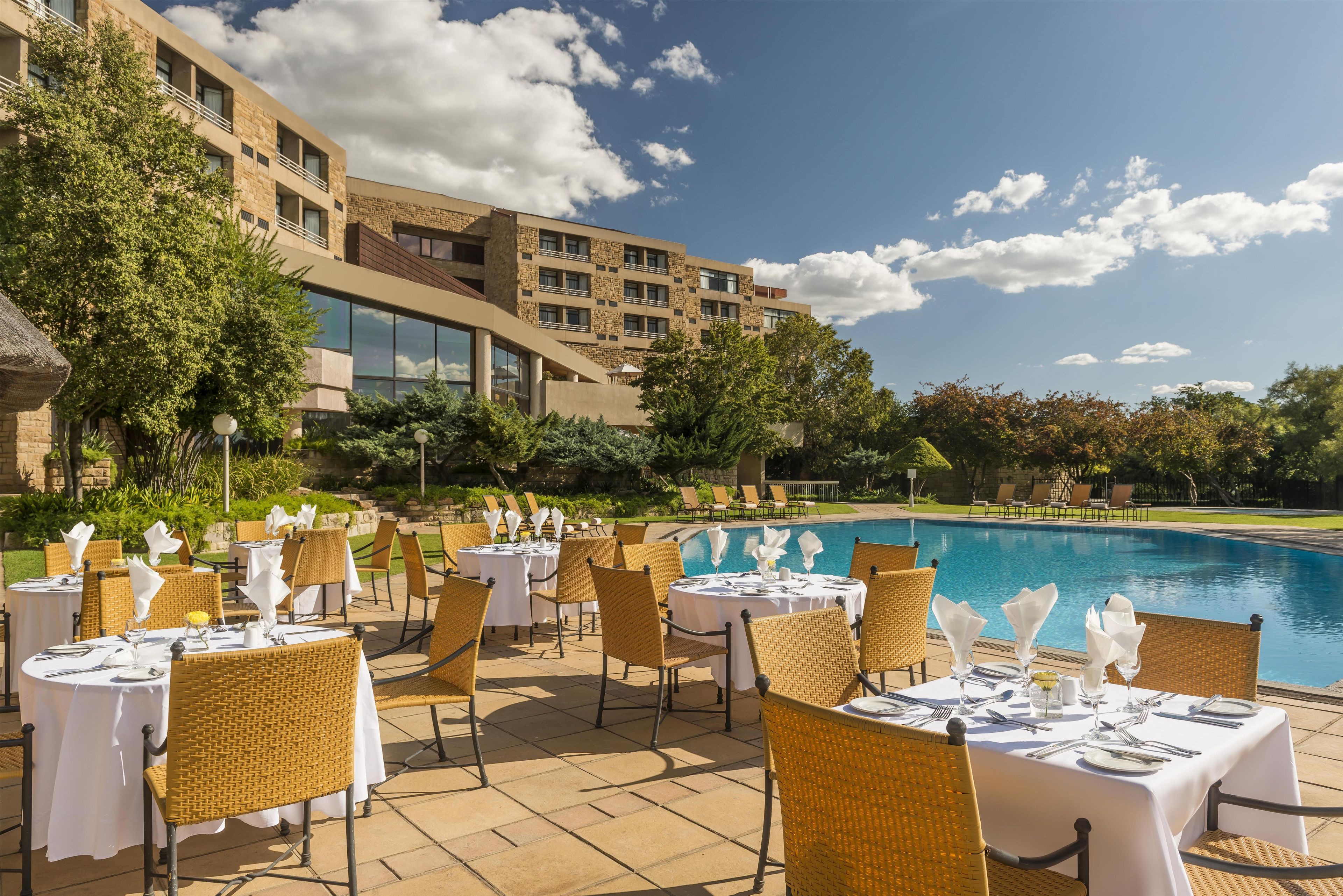 An outdoor dining area with tables set for guests, overlooking a swimming pool, with a building in the background.