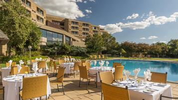 An outdoor dining area with tables set for guests, overlooking a swimming pool, with a building in the background.