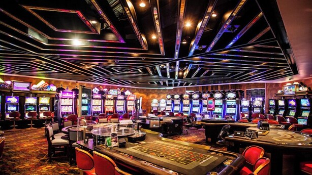 Ein Casino mit mehreren Spielautomaten, einem Roulette-Tisch und roten Barhocker.