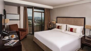A hotel room with a large bed, a desk with a chair, a view of a cityscape through a balcony, and a window with curtains.