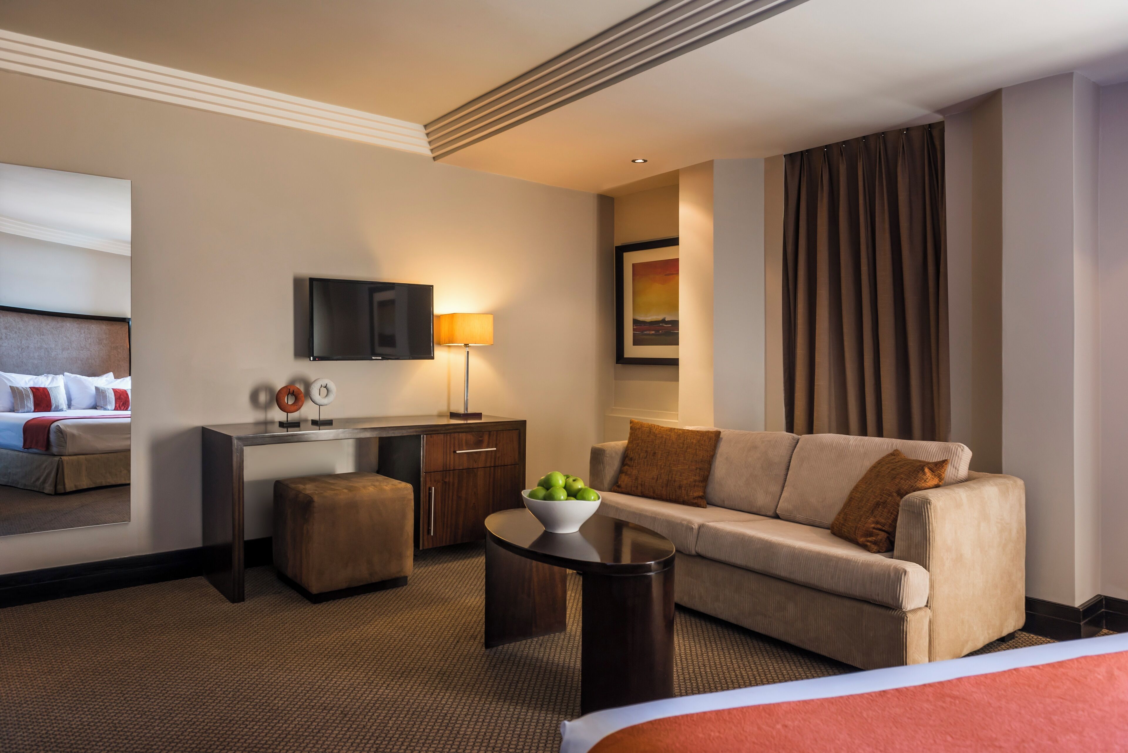 Une chambre d’hôtel avec un lit, un canapé, un bureau, une télévision, une lampe et un panier de fruits.