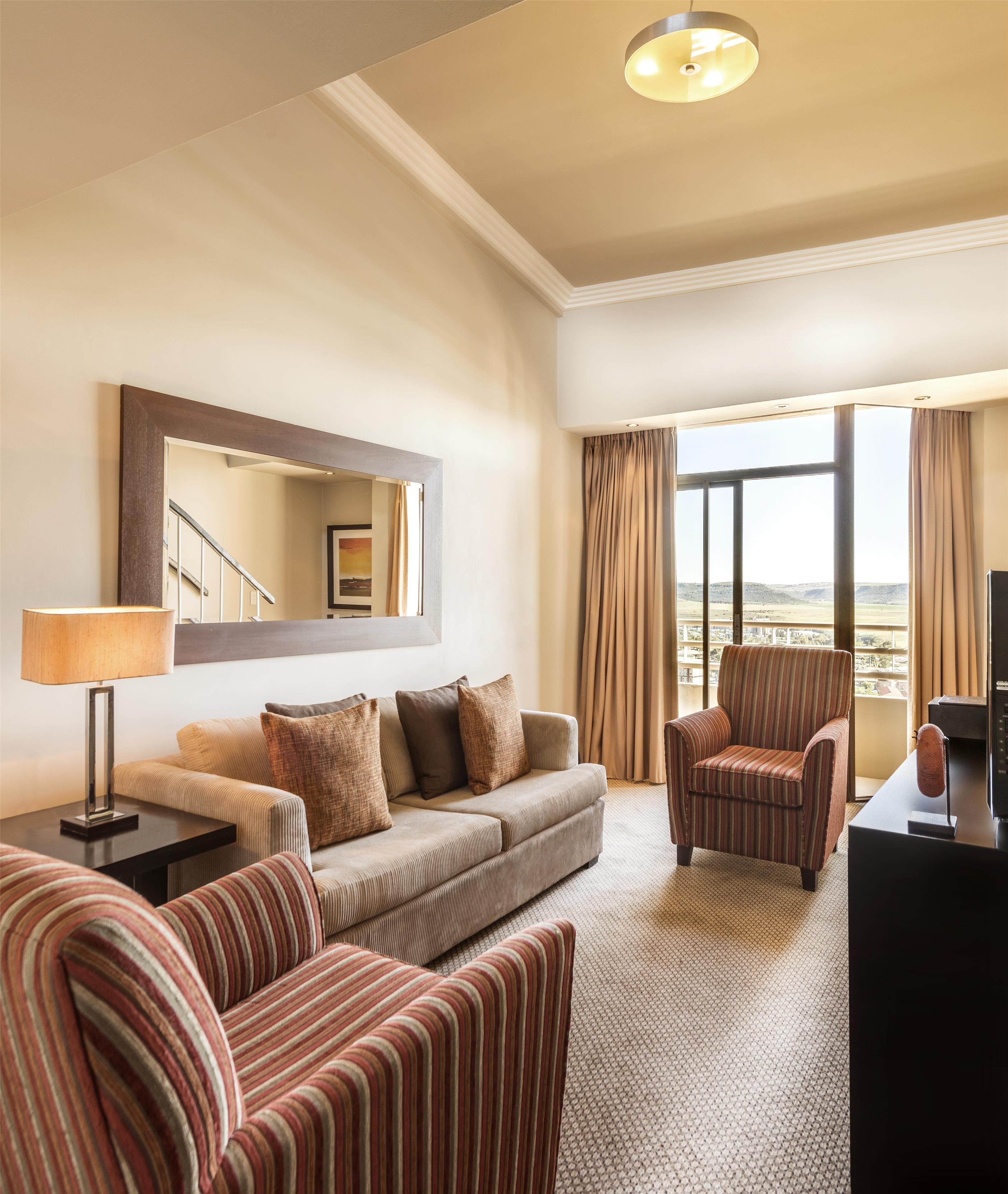 Une chambre d’hôtel équipée d’un canapé, de fauteuils, d’un bureau, d’une lampe, d’une télévision et d’une grande fenêtre avec des rideaux.