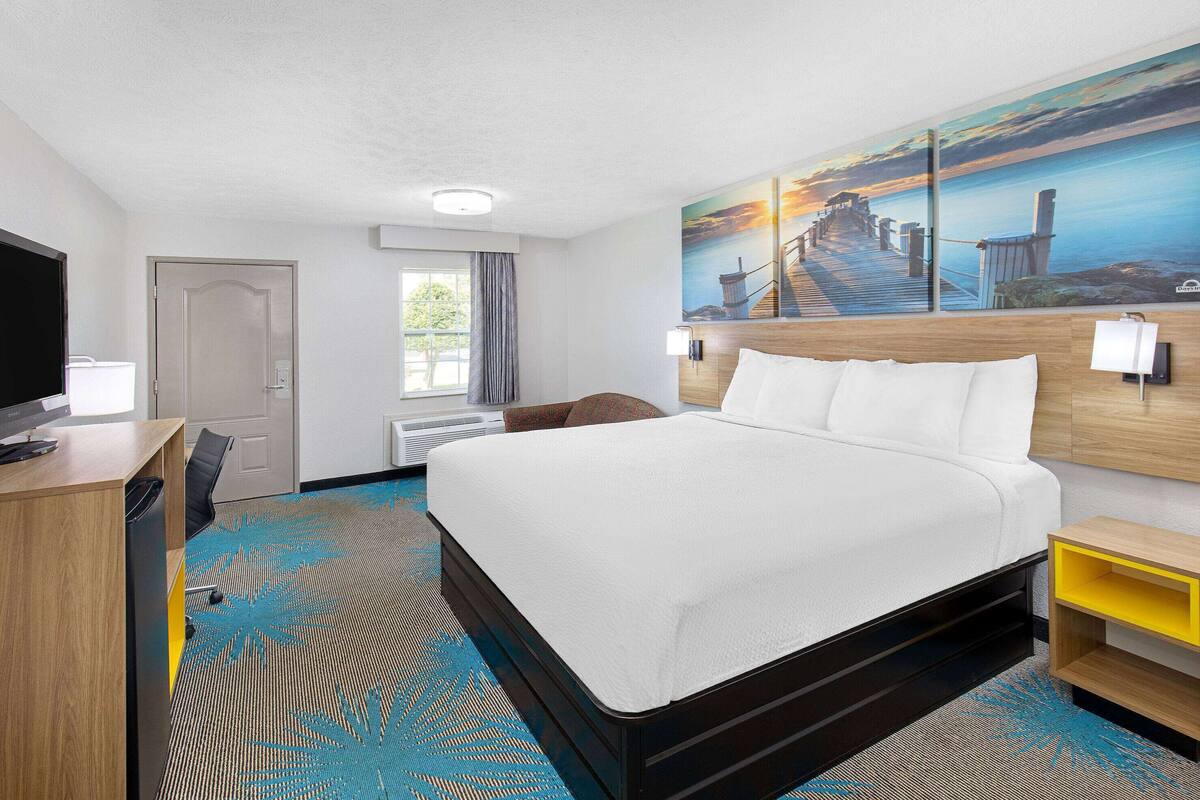 A hotel room with a large bed, a desk with a chair, a TV, a painting on the wall, and a window with curtains.